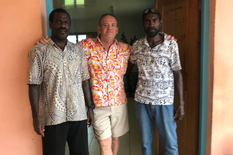 Two ni-Vanuatu men wearing pants and island-themed shirts with an Australian man in the middle wearing an orange floral shirt an