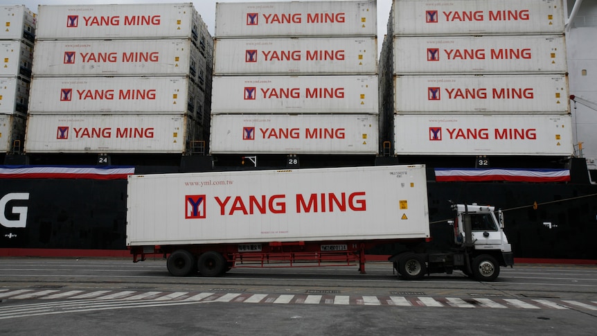 A truck carrying a white shipping container moves past a ship loaded with identical containers stacked four high.