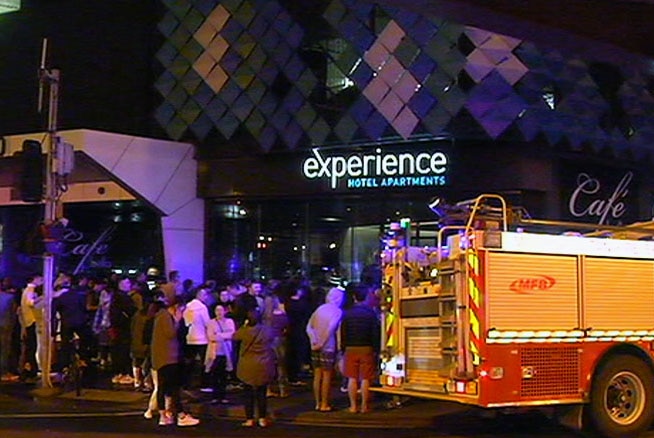 Hotel guests stand outside Experience Bella Hotel Apartments in the dark near a fire truck.