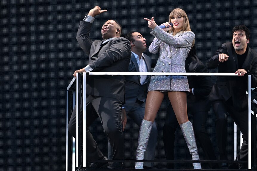 Taylor Swift in a sparkly silver tuxedo jacket, surrounded by back up dancers. 