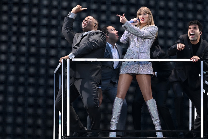 Taylor Swift in a sparkly silver tuxedo jacket, surrounded by back up dancers. 