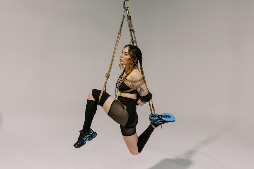 A Taiwanese Australian woman with braided brown and copper hair, is suspended by shibari ropes in black lycra and joggers.