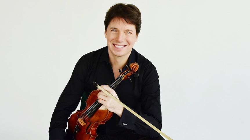 Violinist Joshua Bell sitting on a chair in front of a white background holding his violin and wearing a black shirt