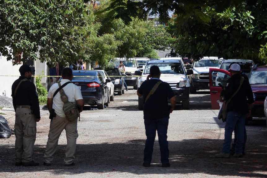 Four men stand in front of police tape with a number of cars in the background. 