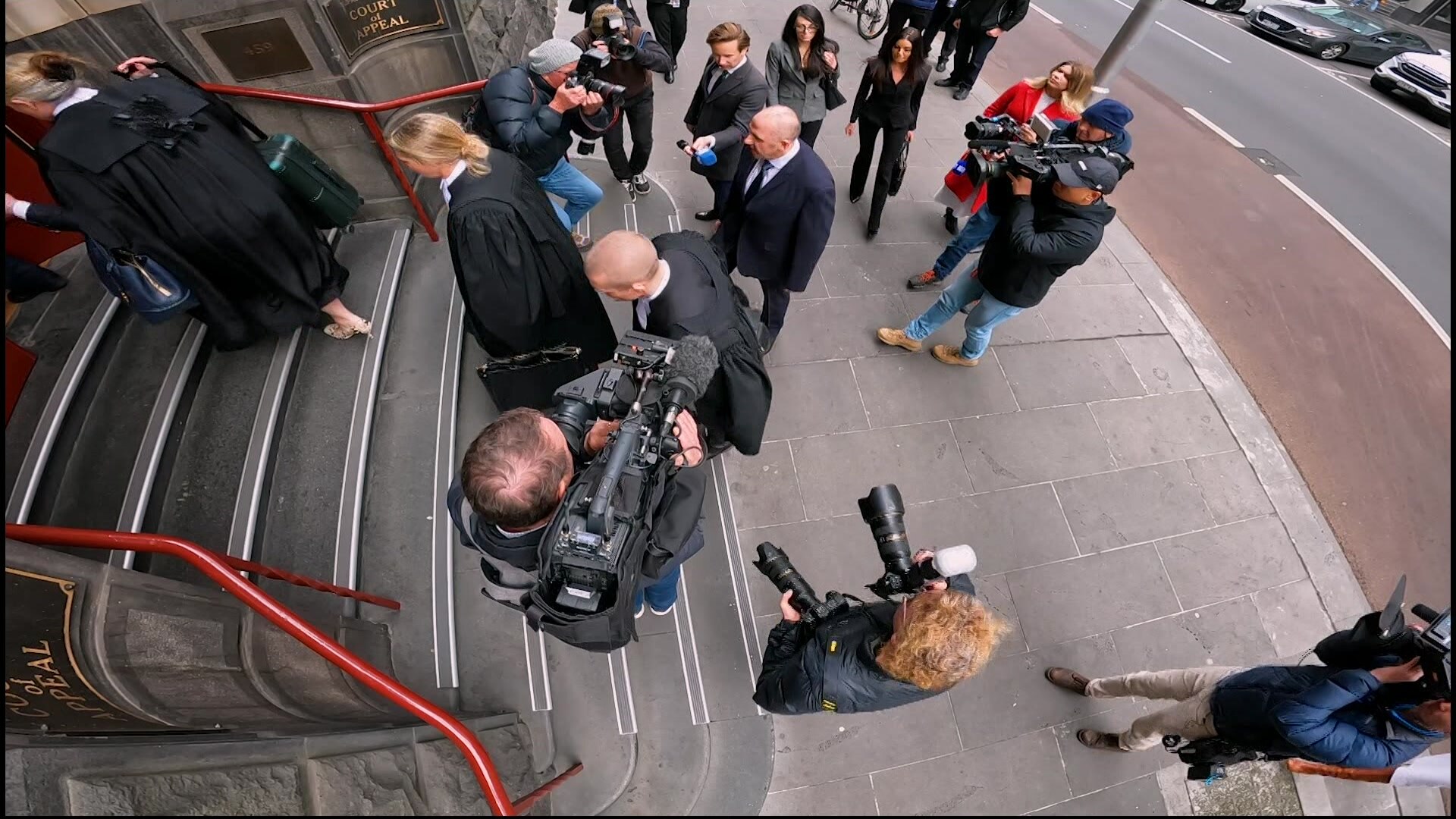 An aerial shot of a bald man in a suit walking up a set of stone steps surrounded by others and tv cameras.