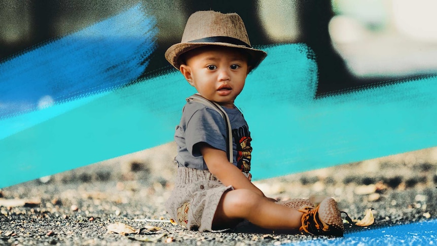 Close up of a siting toddler wearing a fedora with blue brush strokes in the background, for a story about unique baby names.