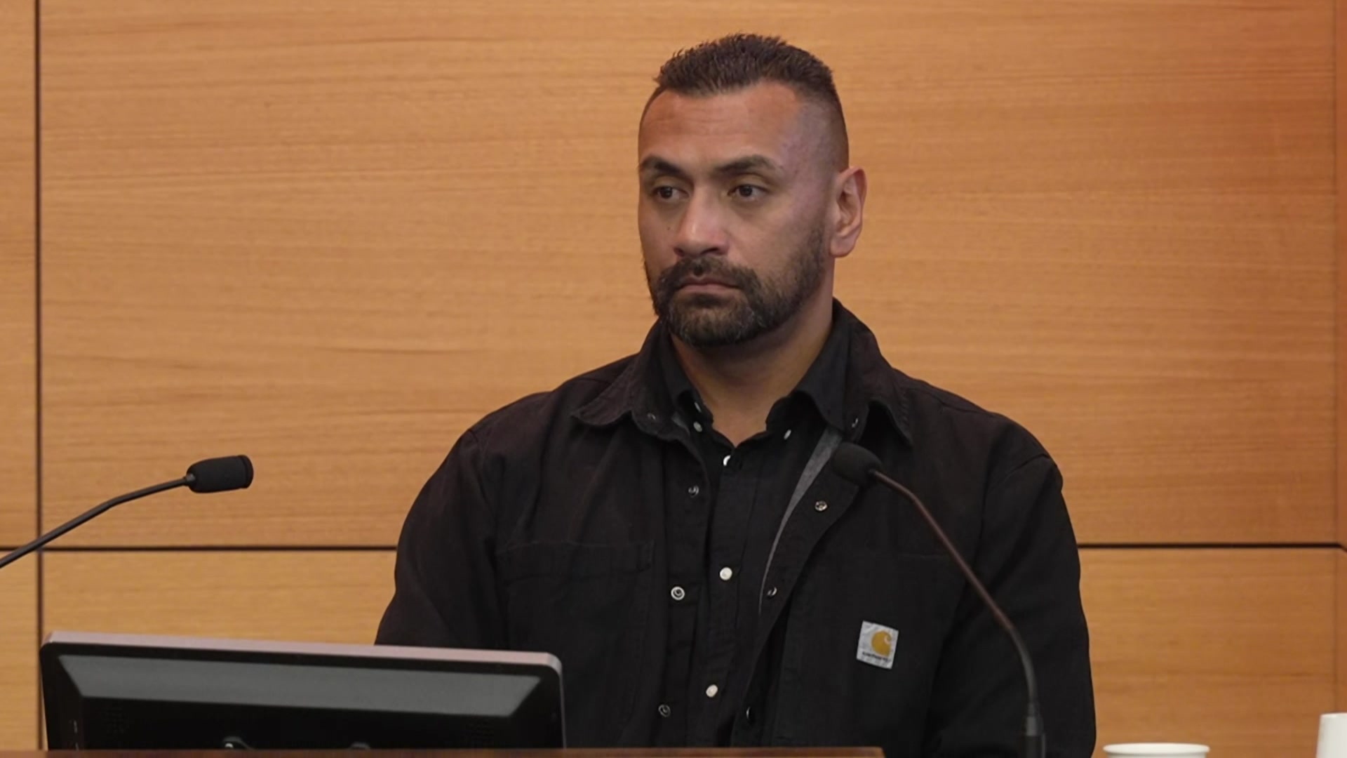 A man in a dark shirt sitting and giving evidence in front of a microphone.