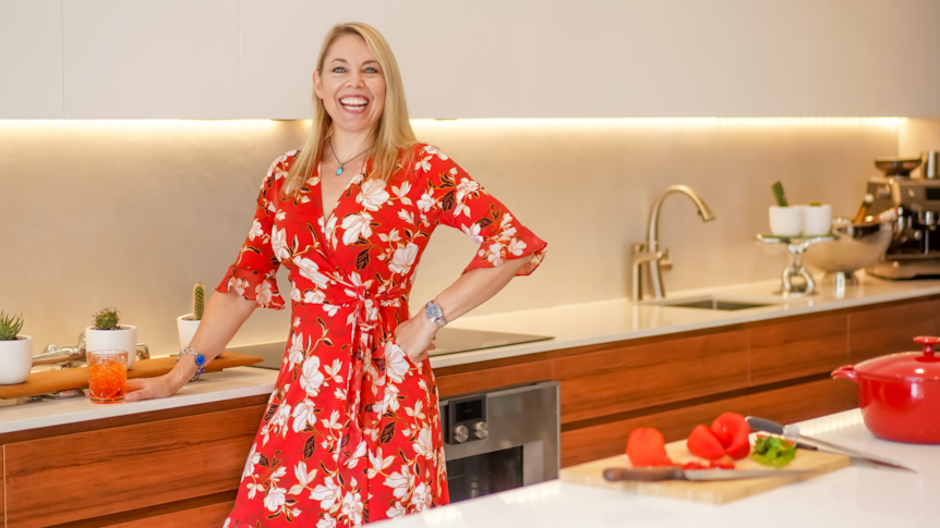 A smiling woman with blonde hair stands in a kitchen with her hand on her hip. 