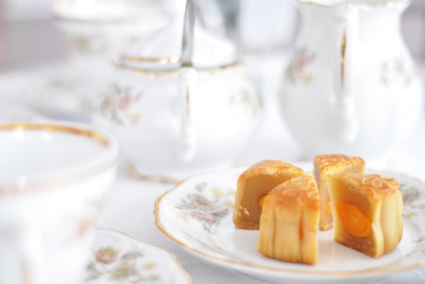 A sliced moon cake in four portions surrounded by tea pots, saucers and cups.