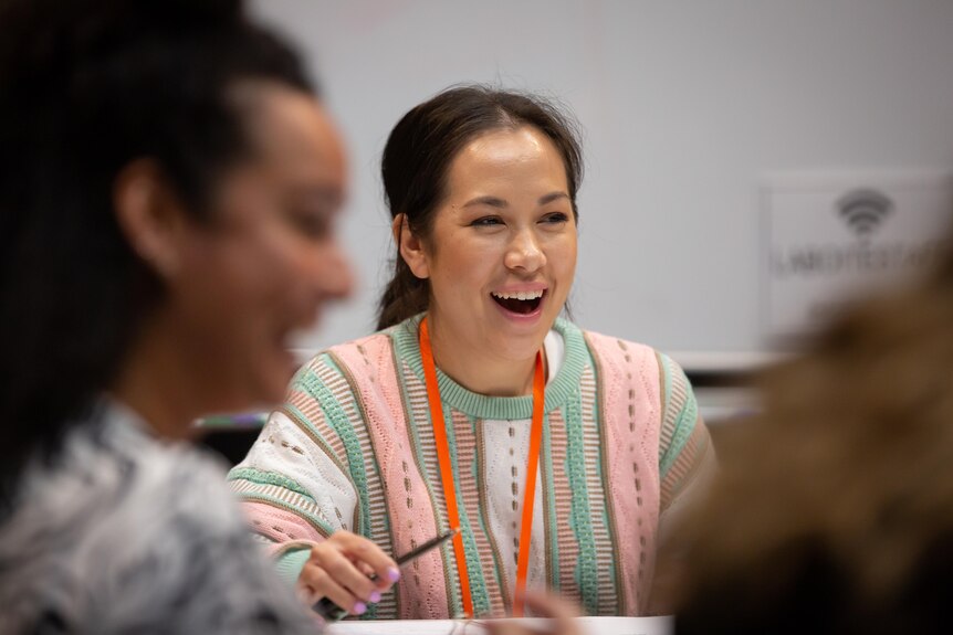 Asian Australian woman with dark hair in a ponytail wears a green and pink striped jumper and sits at a workshop table laughing.