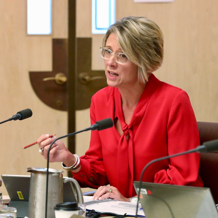 Kristina Keneally asking a question in an Estimates committee. She's wearing a red blouse and clear glasses.