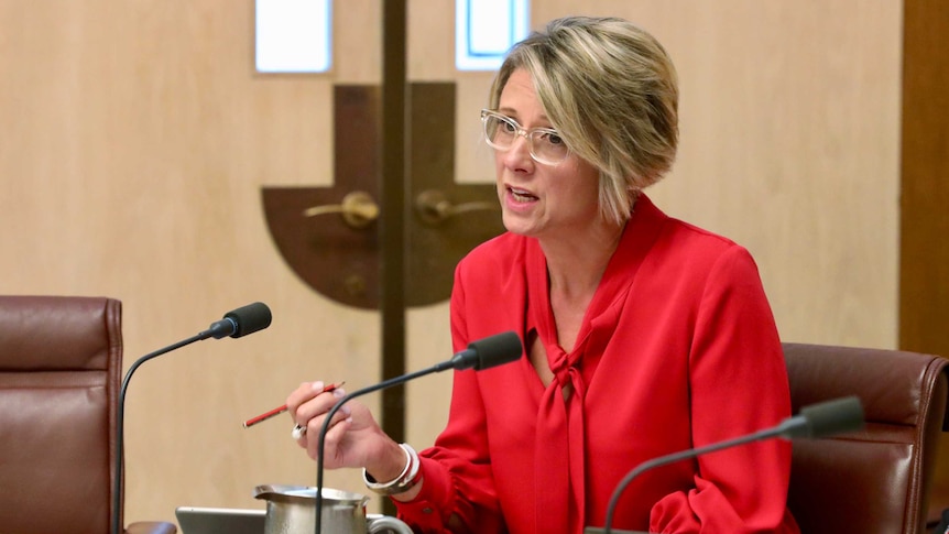 Kristina Keneally asking a question in an Estimates committee. She's wearing a red blouse and clear glasses.