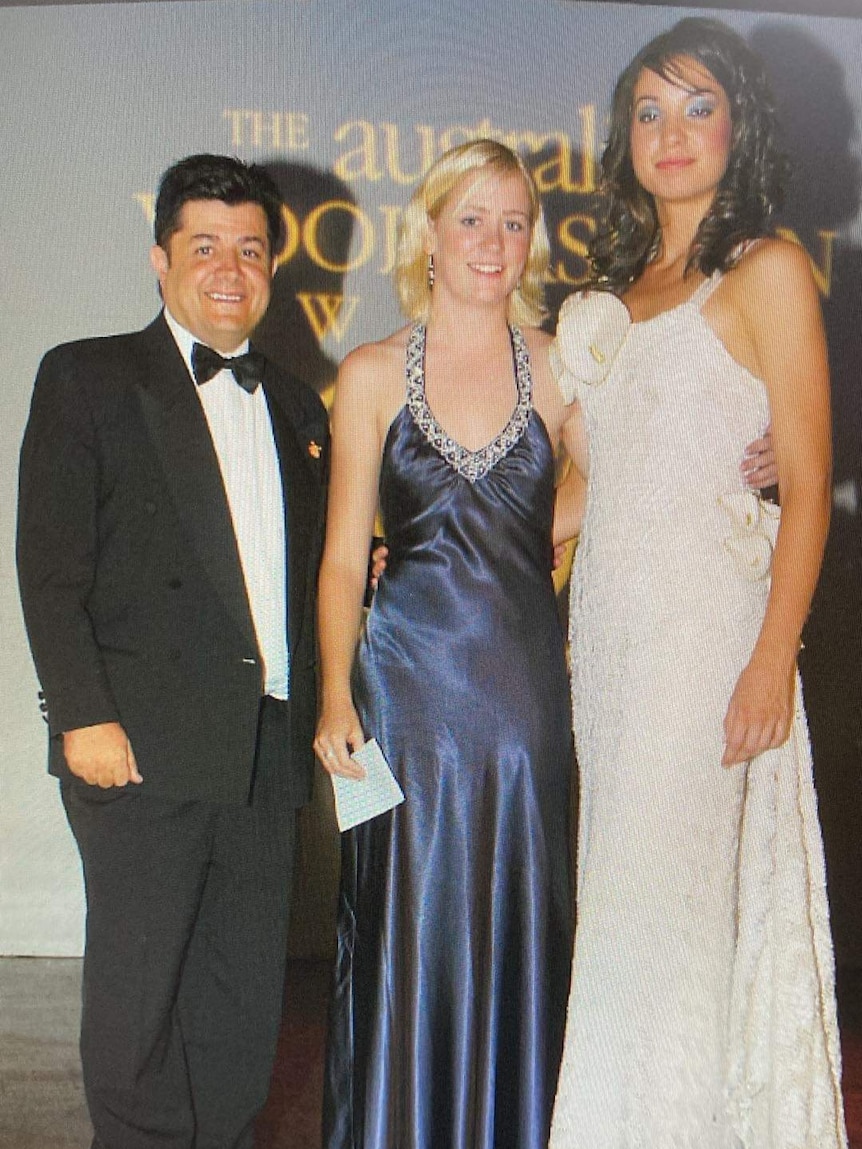 A man, blonde woman, and brunette wearing formal clothing at a ball event and smiling at a the camera