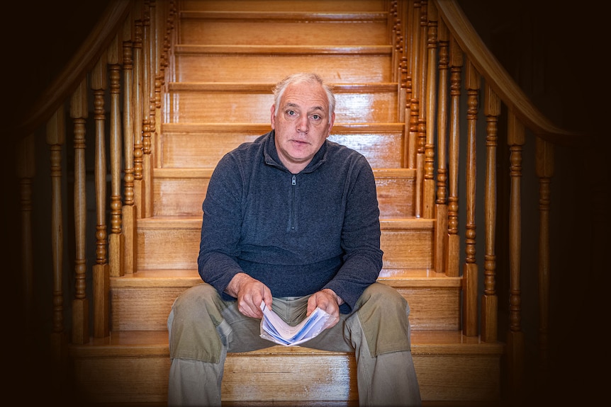 Middle aged man sitting at base of wooden staircase.