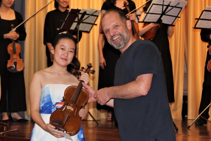 A man hands a violin to a girl standing in front of a string ensemble at a concert performance.