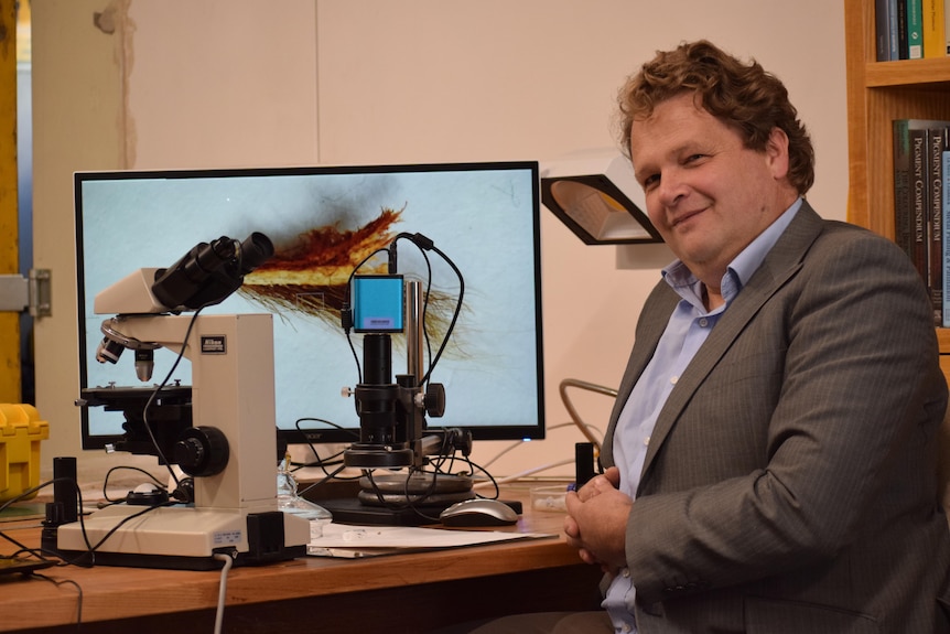 A person in a suit with no tie sits smiling at the camera. They are smiling. Behind them is a microscope and computer screen