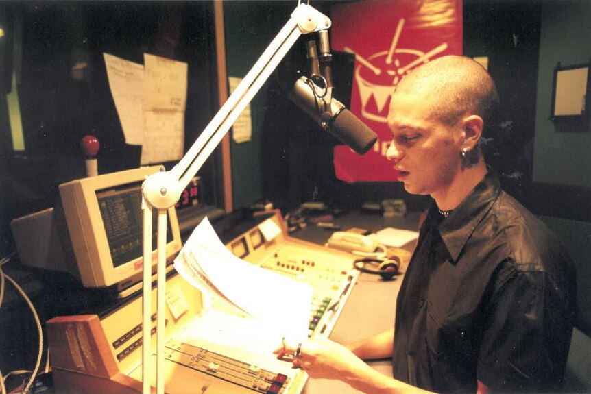 A grainy photo of a young Aboriginal man with a shaved head holding a sheet of paper and speaking into a mic in a studio