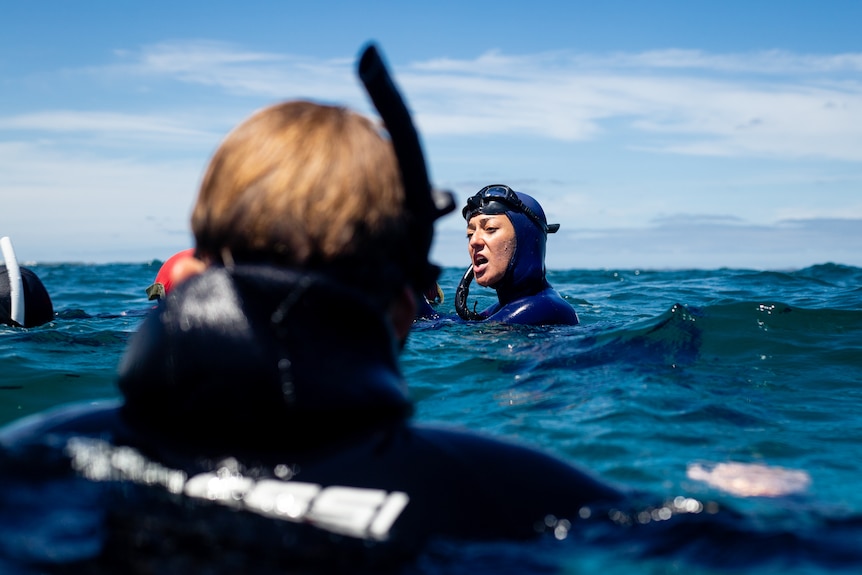A woman in a wetsuit speaks to divers in the open ocean.