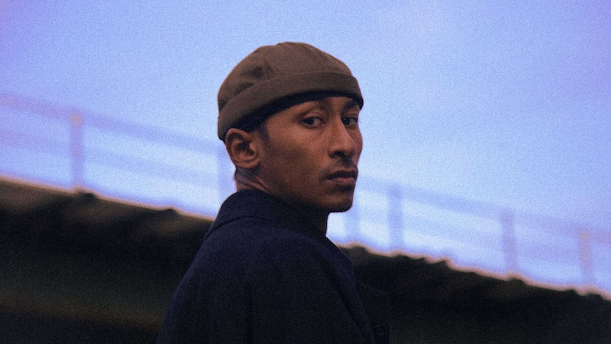 Jitwam wears a brown watch cap & navy jacket. A blue sky at dusk & bridge is in the background.