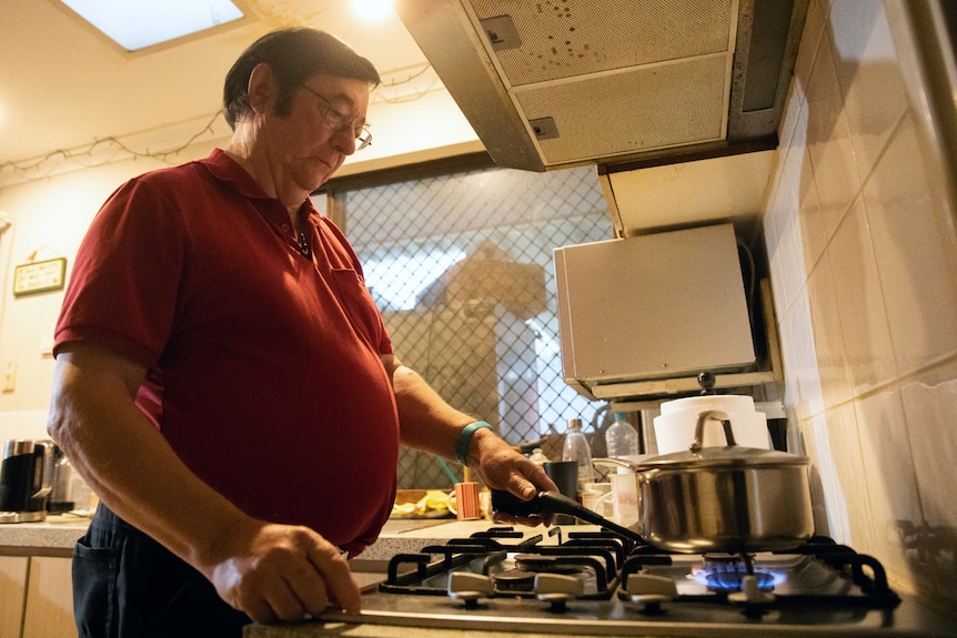 An elderly man in a red shirt cooks in a pot on a gas stove in his kitchen