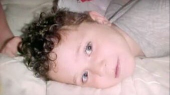 A close-up shot of a toddler with curly hair lying on a mattress with his face turned to the side looking at the camera.