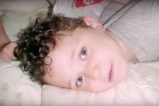 A close-up shot of a toddler with curly hair lying on a mattress with his face turned to the side looking at the camera.