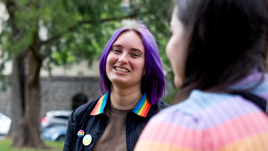 A woman with purple hair and a rainbow collared shirt stands smiling at a person in the foreground.