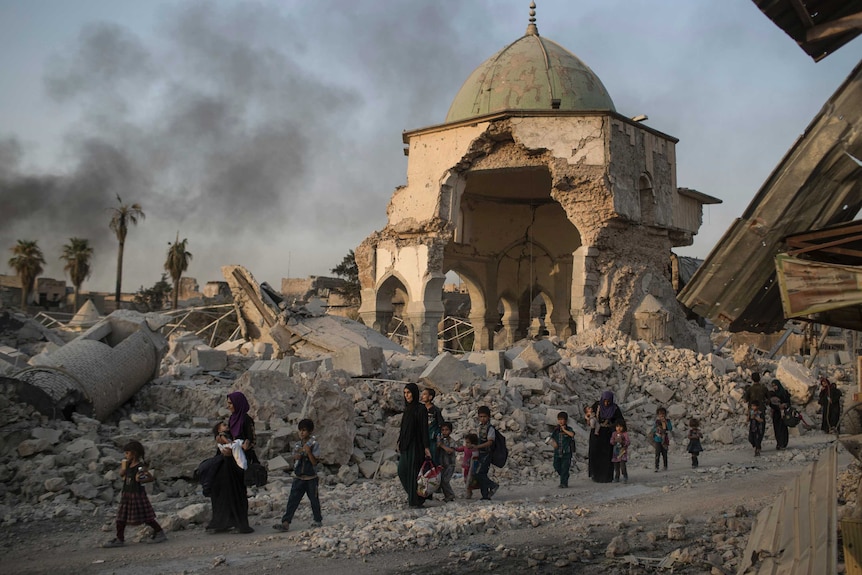 Women and children walk with rubble and a crumbling edifice in the background.