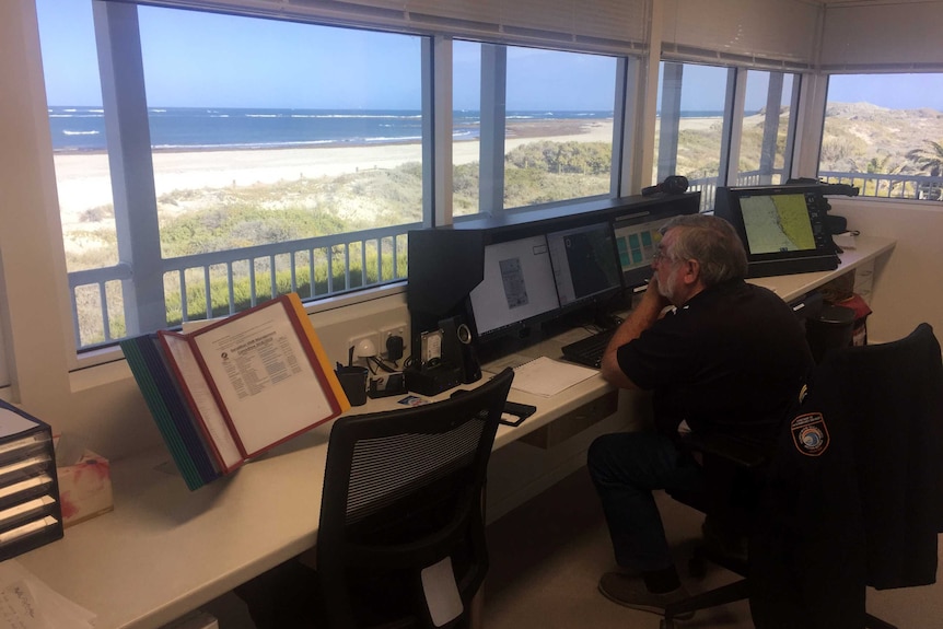 A man sits in a room overlooking a beach in front of computer screens.