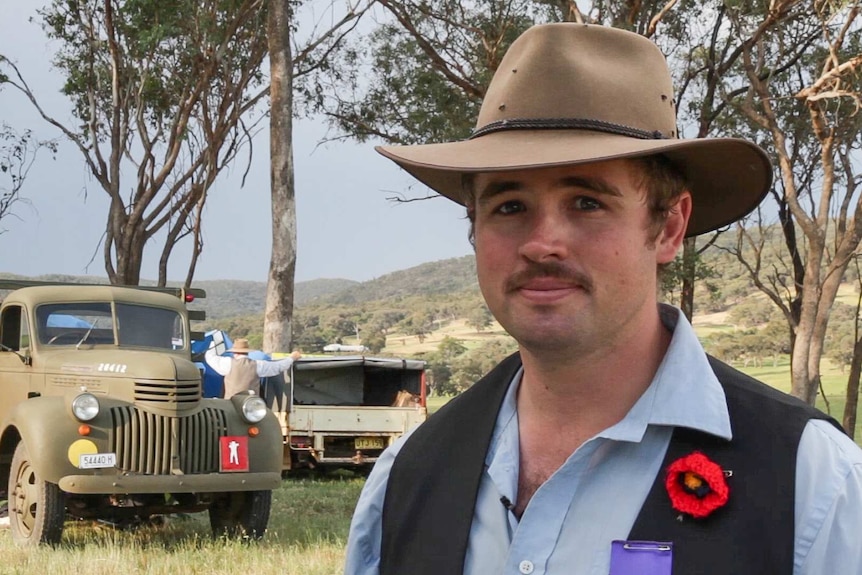 A young man in a hat and WWI period dress and wearing a red poppy stands in front of gum trees and an old army truck