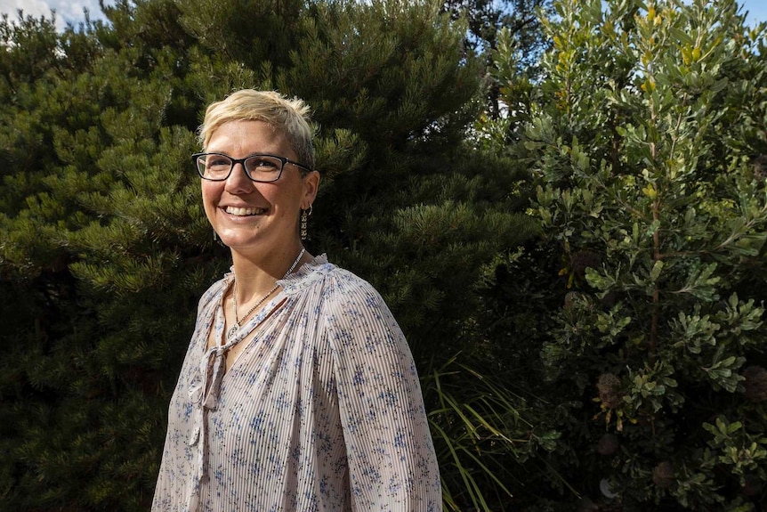 Une femme aux cheveux blonds courts et lunettes debout dans un jardin sourit à la caméra.