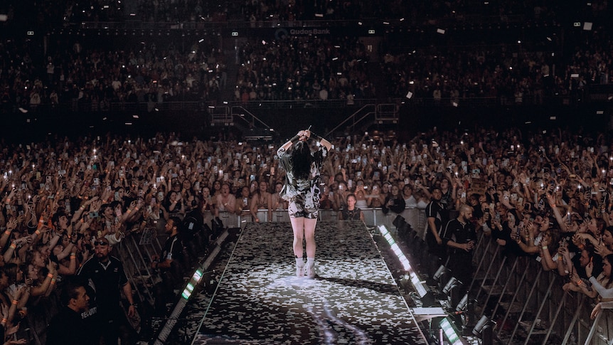Billie Eilish stands at the end of a catwalk overlooking a packed arena audience