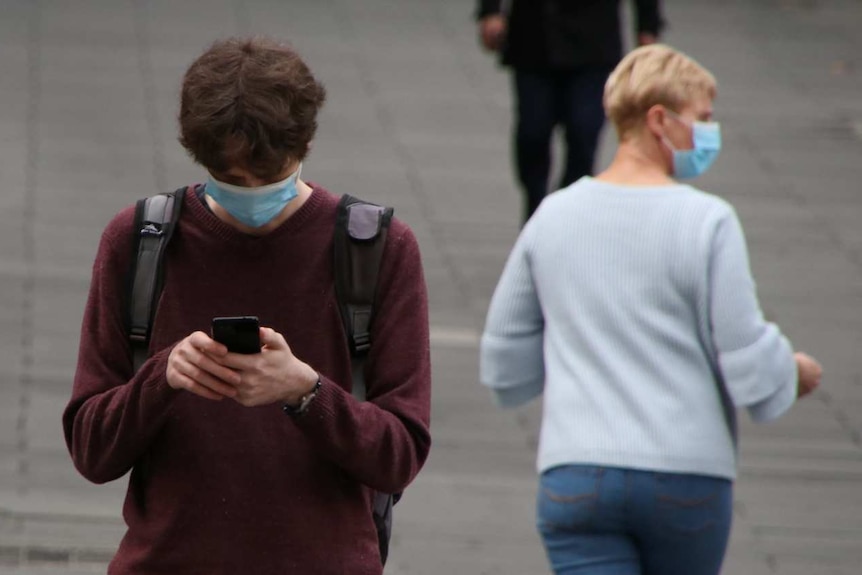 On a city street, a person wearing a face mask looks down at their phone, while a woman in a mask can be seen in the background.