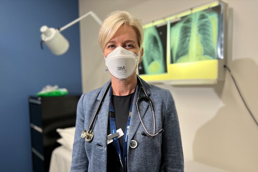 A woman wearing a stethoscope and mask stands in a hospital room.