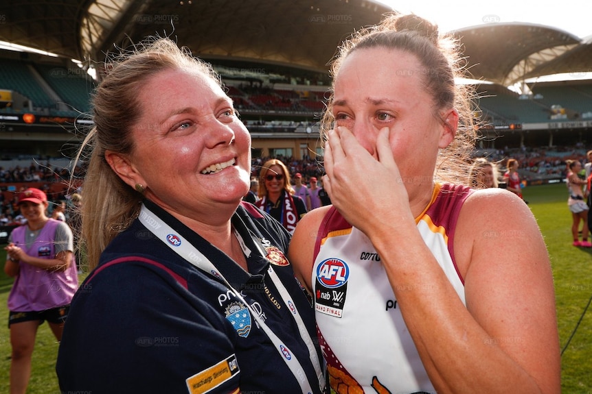 Two women celebrate on a football field, one wearing a uniform and the other a polo shirt