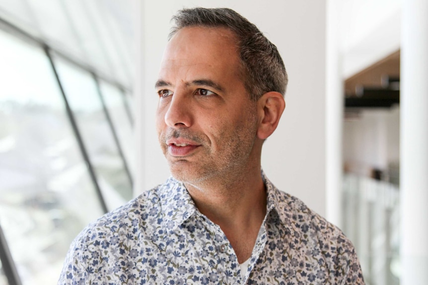 Man with greying hair and floral shirt stands in a corridor, looking towards a window.