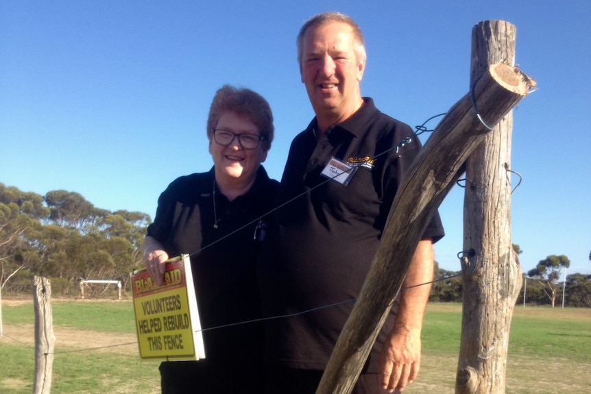 A man and a woman wearing black, standing in front of fencing poles and a roll of wire.