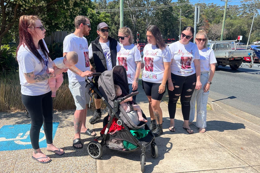 A group of people in matching T-shirts stand on a street corner, looking sad.