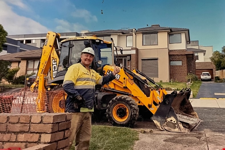 A man wearing construction fluros stands in front of a digger giving a thumbs-up.