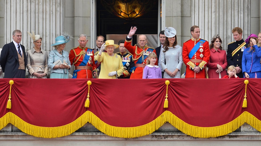 The royal family, all decked out in finery, including men in military uniform, stand on a balcony and wave