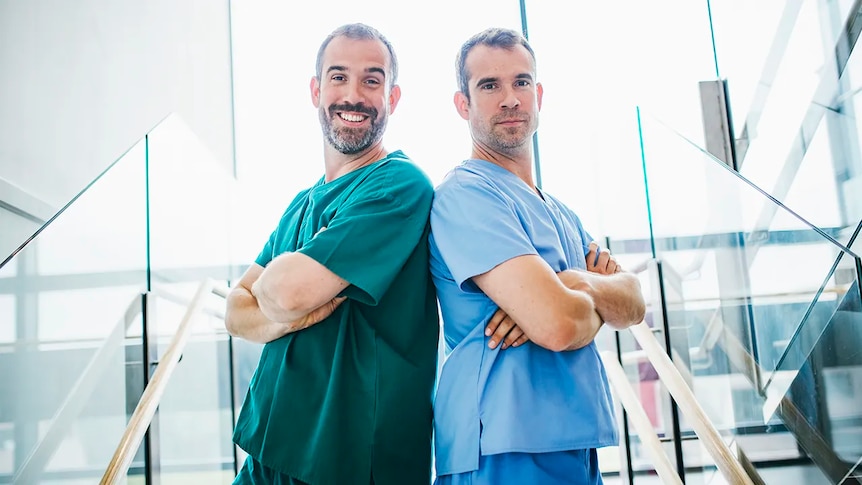 Twin men are smiling back to back with arms crossed wearing hospital scrubs