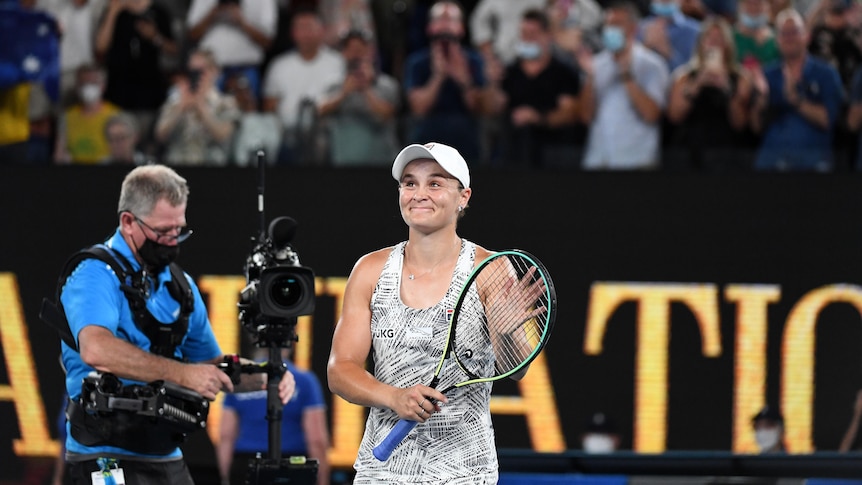 Ash Barty claps her hand to her racquet to acknowledge the crowd after an Australian Open win. A camera man walks behind her.