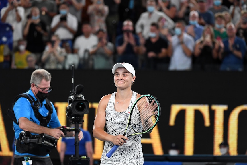 Ash Barty claps her hand to her racquet to acknowledge the crowd after an Australian Open win. A camera man walks behind her.