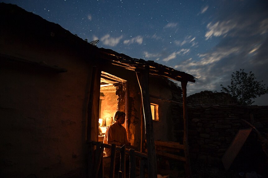 A woman backlit by warm interior light sits at doorway of countryside shack and looks at at night sky.