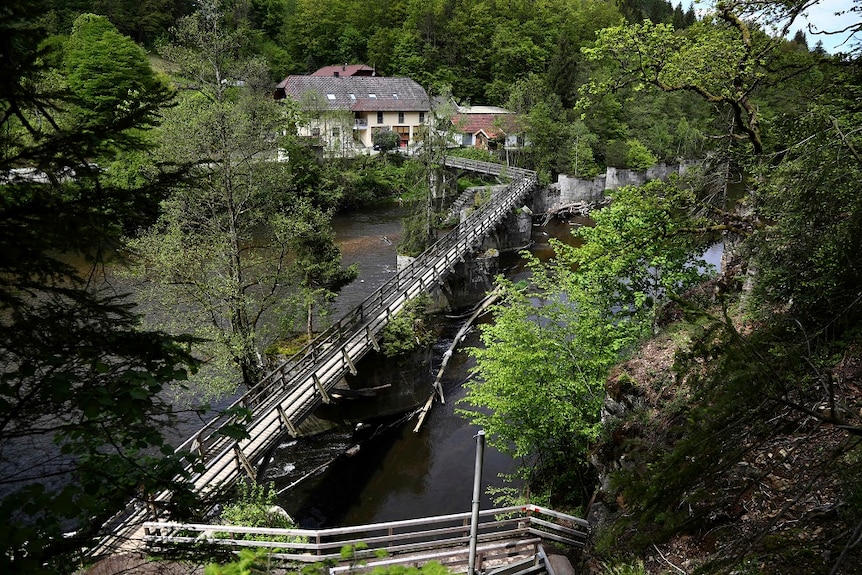 A walkway over a river leads to a hotel sitting on the river bank in front of a verdant forest.