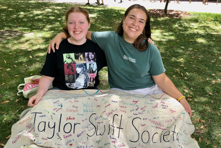 Two women smiling and holding each other's shoulders in front of a sign that reads "Taylor Swift Society".