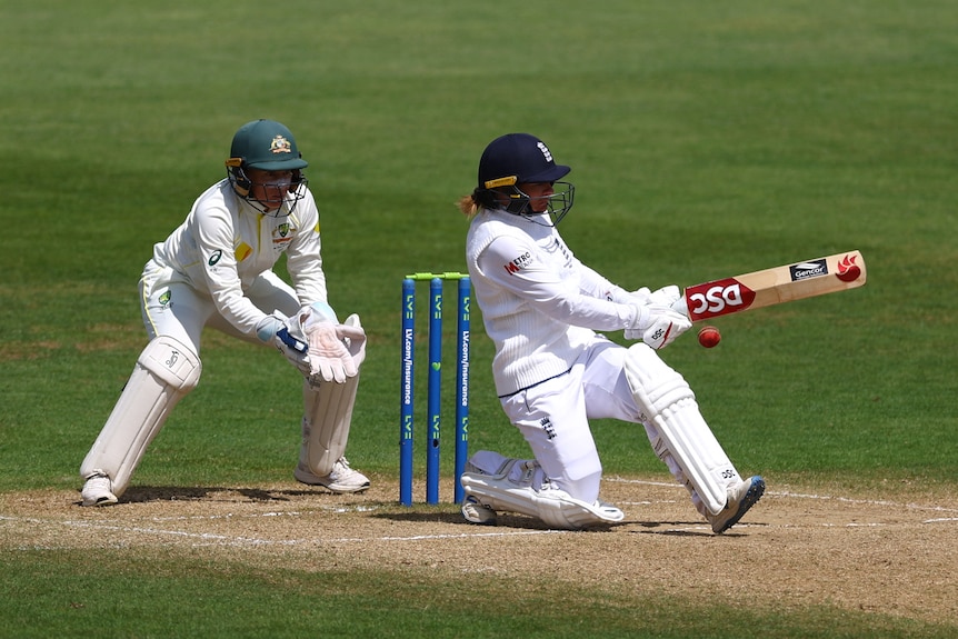 A woman behind a wicket with her hands placed ready to catch a ball while a woman batting hits a cricket ball. 