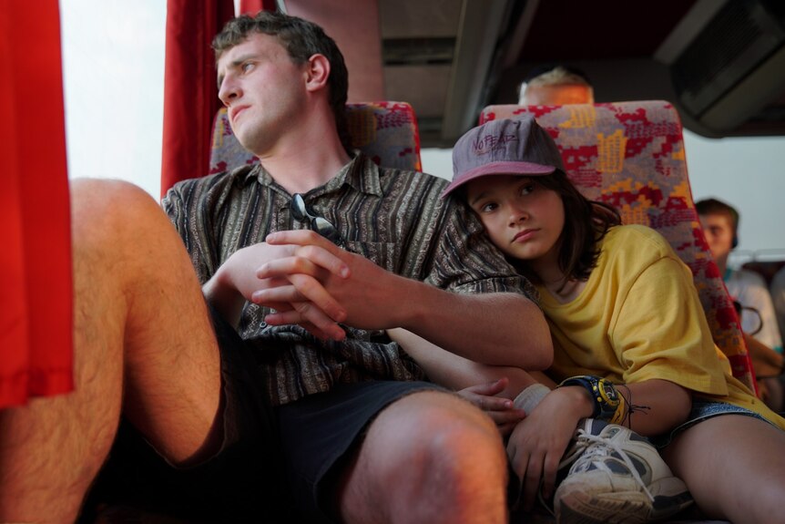 A 12 year old girl leaning on a man in his late 20s - both in early 2000s clothes - on a bus