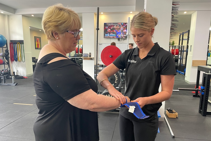 A woman applies a wrist strap to another woman's arm at the gym.