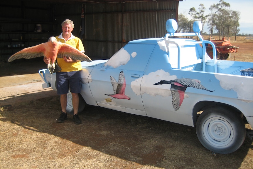 A man in black shorts and a yellow shirt stands next to a ute painted light blue with pink galahs on it.